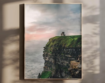 May include: A vertical wall art print featuring a landscape photograph of a stone tower perched atop a steep, grassy cliff overlooking the ocean. The sky displays soft pink and blue hues during twilight, contrasting with the dark, rugged rock face.