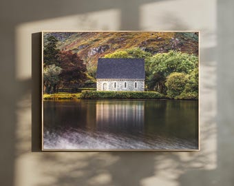 Peut inclure: Une impression d'art mural rectangulaire représentant un bâtiment en pierre avec un toit en pente gris foncé situé au bord d'un lac calme. La scène comprend des arbres verts luxuriants et une colline en arrière-plan, avec le reflet du bâtiment.