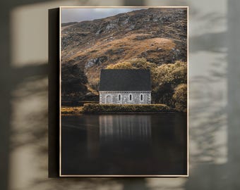 Peut inclure: Une impression d'art mural encadrée représentant un bâtiment en pierre au toit sombre situé au bord d'un lac calme. Derrière la structure, une grande pente de montagne accidentée s'élève avec des taches de végétation brune et verte sous un ciel nuageux.