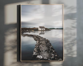 Peut inclure: Une impression d'art mural rectangulaire présentant une photographie de paysage d'un petit cottage blanc situé au bord d'un lac calme. Un chemin en pierre mène vers le bâtiment sous un ciel nuageux, le tout encadré par un fin cadre en bois clair.