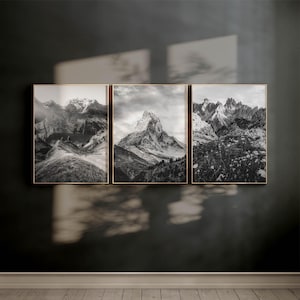 Puede incluir: Un tríptico de fotografías en blanco y negro enmarcadas de paisajes de montaña. Cada impresión enmarcada presenta una escena de montaña diferente, incluyendo picos nevados y valles. Los marcos son de color dorado cálido y la obra de arte se exhibe en una pared oscura.