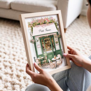 May include: A framed print of a watercolor illustration of a Parisian cafe with a pink awning that says "Petit Amour". The cafe has green doors and windows and is decorated with flowers. There are tables and chairs outside the cafe.