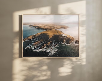 May include: An aerial photograph of a rocky coastline with a lighthouse on a cliff. The ocean is a deep blue and the sky is a light blue with white clouds. The sun is shining brightly and the water is sparkling.