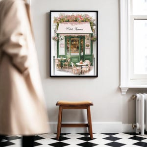 May include: A watercolor painting of a Parisian cafe with a green awning that says "Petit Amour". The cafe has tables and chairs outside and is surrounded by flowers.