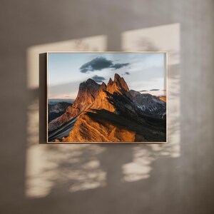 Puede incluir: Una fotografía de una cordillera con un brillo dorado al atardecer. Los picos son afilados y dentados, y el cielo es de un azul suave con algunas nubes.