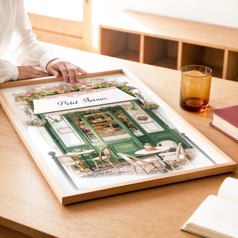 May include: A watercolor painting of a Parisian cafe called "Petit Amour". The cafe has a green door and awning, and there are tables and chairs set up outside. The painting is framed in a light wood frame.