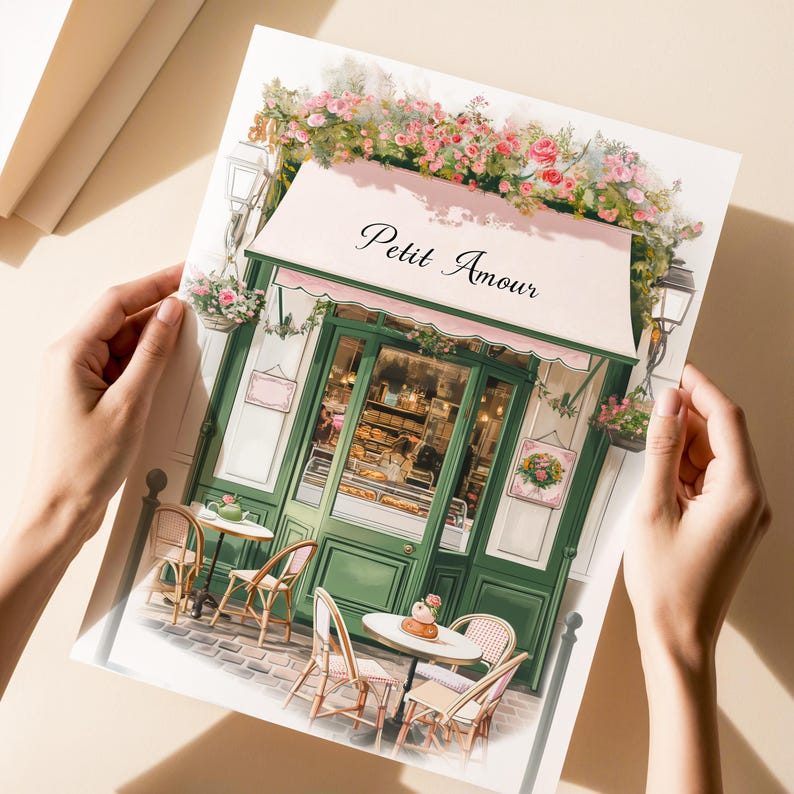 May include: A watercolor illustration of a Parisian cafe with a pink awning that reads "Petit Amour". The cafe has green doors and windows, and there are tables and chairs set up outside. There are flowers and greenery around the cafe.