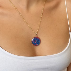 May include: A gold-colored necklace with a round pendant. The pendant features a red border and a blue and red design. The necklace is worn on a person wearing a white tank top.