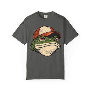 Pode incluir: Uma t-shirt de manga curta cinzento-carvão com uma ilustração gráfica de um sapo verde a usar um boné de basebol laranja e branco. O sapo tem uma expressão severa com contornos pretos detalhados sobre o tecido cinzento escuro da camisa.