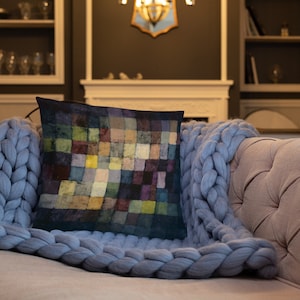 May include: A square throw pillow featuring a grid pattern of small, multicolored squares in shades of deep blue, yellow, green, and purple. The pillow is resting on a light blue, chunky knit blanket that is draped over a tufted sofa.