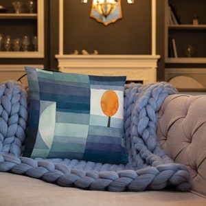 May include: A throw pillow with a blue and green abstract design featuring a single orange tree. The pillow is on a blue knitted throw blanket.