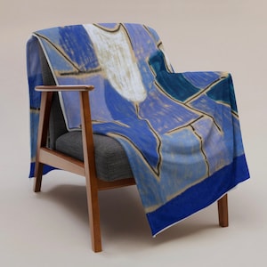 May include: A soft throw blanket featuring an abstract pattern of blue, light blue, and white geometric shapes with dark outlines, draped over a dark gray armchair with wooden armrests and legs against a neutral background.