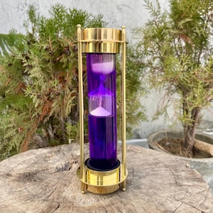Puede incluir: Un reloj de arena decorativo con líquido morado y arena blanca. El reloj de arena está encerrado en un marco de metal dorado. El reloj de arena mide aproximadamente 15 cm de alto.