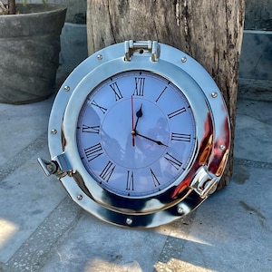 Może przedstawiać: Okrągły, srebrny zegar ścienny z nautycznym wzorem okna okrętowego. Tarcza zegara jest biała z czarnymi rzymskimi cyframi i czarnymi wskazówkami. Zegar jest umieszczony na tle zwietrzałego drewna.