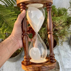 Może przedstawiać: Drewniana klepsydra z ciemnobrązowym wykończeniem. Klepsydra ma przezroczystą szklaną komorę wypełnioną białym piaskiem. Klepsydra jest wspierana przez drewnianą ramę z dekoracyjnymi toczonymi kolumnami.