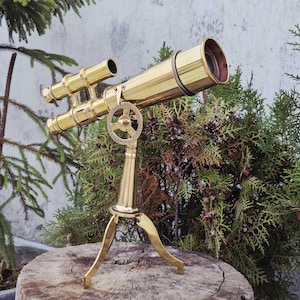 May include: A brass telescope with a tripod base. The telescope has a long barrel with a lens at one end and an eyepiece at the other. It features a small, secondary scope and a circular adjustment dial. The telescope is displayed on a wooden stump.