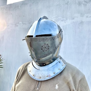 May include: A polished silver knight's helmet with a visor and decorative floral patterns. The helmet features a rounded top and a neck guard with rivets. The person is wearing a tan corduroy shirt.