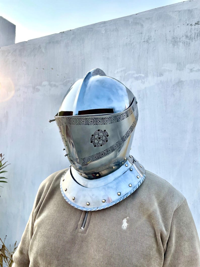May include: A polished silver knight's helmet with a visor and decorative floral patterns. The helmet features a rounded top and a neck guard with rivets. The person is wearing a tan corduroy shirt.