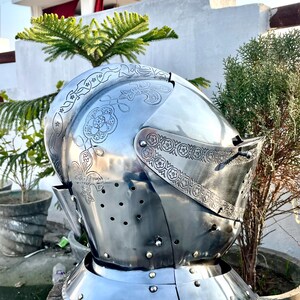 May include: A polished steel medieval helmet with decorative floral and scrollwork engravings. The helmet features a visor with small eye slits and numerous ventilation holes. The neck guard is riveted to the helmet's base. The helmet is displayed on a wooden stump.