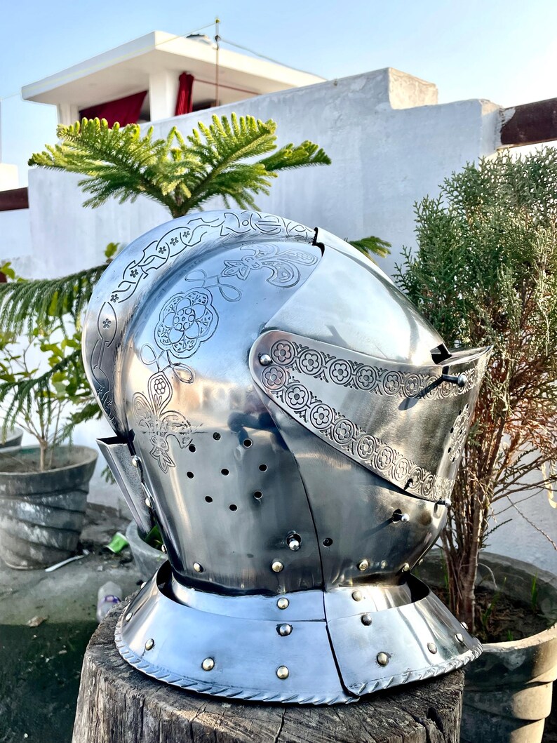 May include: A polished steel medieval helmet with decorative floral and scrollwork engravings. The helmet features a visor with small eye slits and numerous ventilation holes. The neck guard is riveted to the helmet's base. The helmet is displayed on a wooden stump.