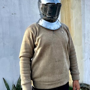 May include: A medieval-style helmet with a visor and neck guard. The helmet is made of polished metal and has a dark visor. The neck guard is a separate piece with rivets. The person is wearing a tan corduroy sweater and dark blue jeans.