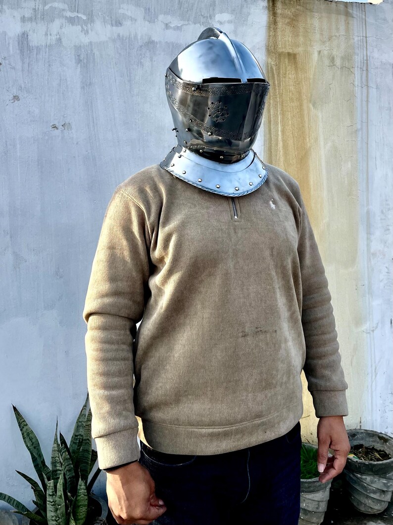May include: A medieval-style helmet with a visor and neck guard. The helmet is made of polished metal and has a dark visor. The neck guard is a separate piece with rivets. The person is wearing a tan corduroy sweater and dark blue jeans.