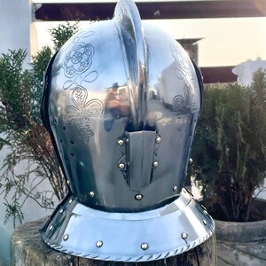 May include: A polished steel helmet with floral engravings. The helmet features a prominent crest and a neck guard with rivets. The surface reflects the surrounding environment, and the helmet is displayed on a wooden surface.