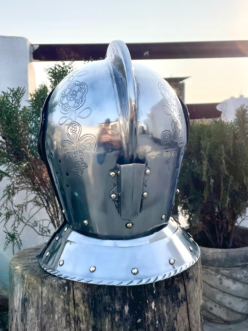 May include: A polished steel helmet with floral engravings. The helmet features a prominent crest and a neck guard with rivets. The surface reflects the surrounding environment, and the helmet is displayed on a wooden surface.