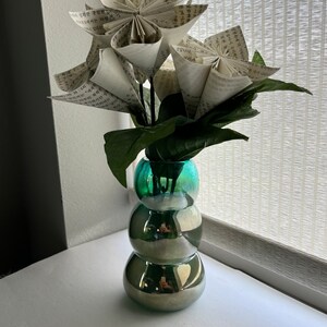 May include: A bouquet of paper flowers made from book pages in a teal blue vase with a textured surface. The vase has a round, bulbous shape.