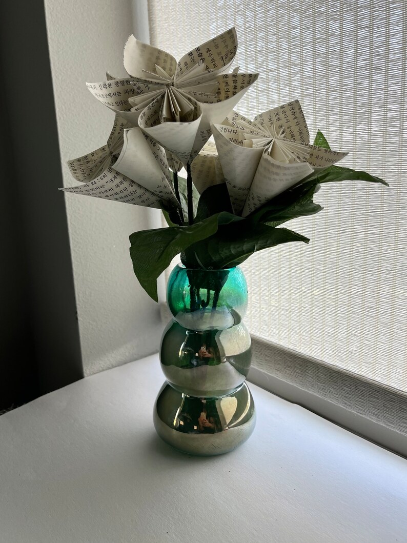 May include: A bouquet of paper flowers made from book pages in a teal blue vase with a textured surface. The vase has a round, bulbous shape.