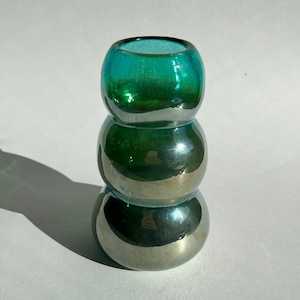 May include: A teal green glass vase with a rounded, bulbous shape. The vase is made of iridescent glass, giving it a shimmering effect.