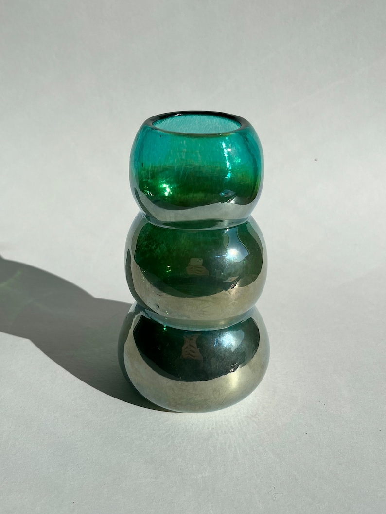 May include: A teal green glass vase with a rounded, bulbous shape. The vase is made of iridescent glass, giving it a shimmering effect.