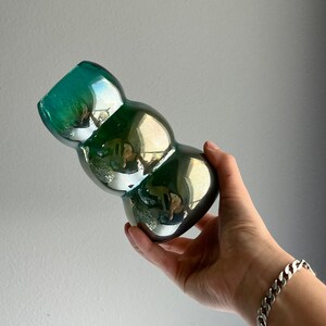 May include: A green iridescent glass vase with a bulbous, rounded shape. The vase is made of thick glass and has a smooth, shiny finish.