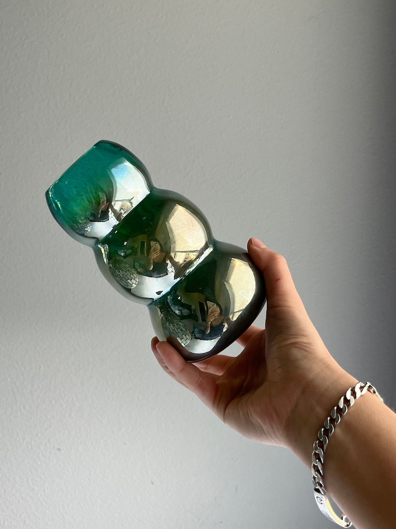 May include: A green iridescent glass vase with a bulbous, rounded shape. The vase is made of thick glass and has a smooth, shiny finish.
