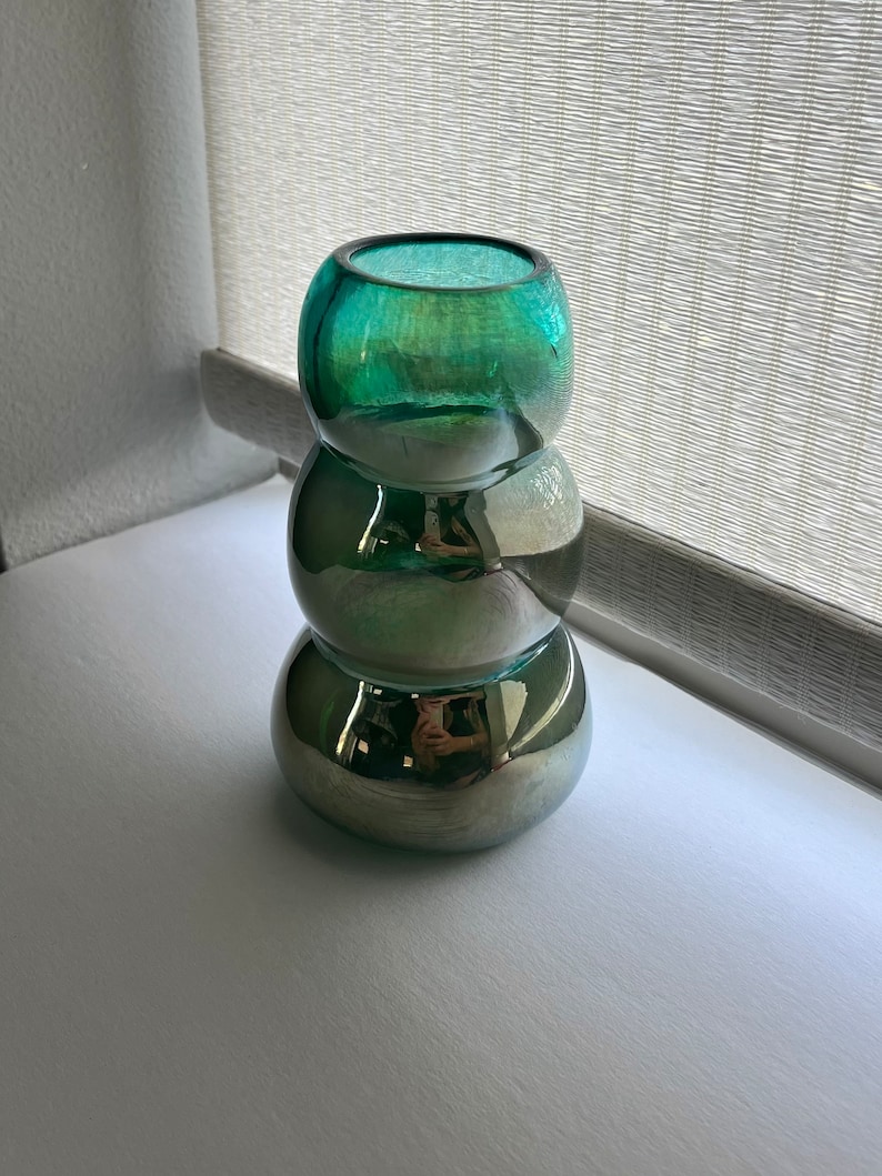May include: A stack of three round glass vases in shades of green and silver. The vases are stacked on top of each other, with the largest vase at the bottom and the smallest vase at the top.
