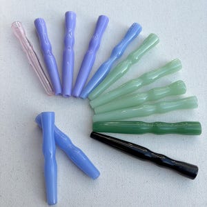 Puede incluir: Una colección de accesorios para fumar de vidrio de colores en varios tonos, incluyendo rosa, lavanda, azul, verde y negro. Las piezas de vidrio tienen un diseño acanalado y están dispuestas sobre una superficie blanca.