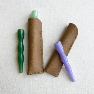 May include: An overhead shot shows two glass pipes, one green and one purple, next to two brown leather cases with green stitching. The pipes have a unique, curved design. The cases are open, revealing the tops of the pipes.