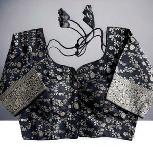 May include: A black brocade blouse with a floral pattern and silver thread embroidery. The blouse has a scoop neck and short sleeves. It features a hook and eye closure at the front and a tie-up back.