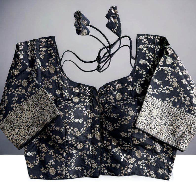 May include: A black brocade blouse with a floral pattern and silver thread embroidery. The blouse has a scoop neck and short sleeves. It features a hook and eye closure at the front and a tie-up back.