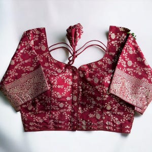 May include: A maroon brocade blouse with a floral pattern and silver accents. The blouse has a scoop neck and short sleeves. The back of the blouse has a tie closure.