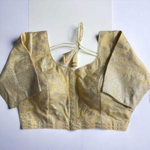 May include: A pale yellow brocade blouse with a silver floral pattern and a tie closure at the back.