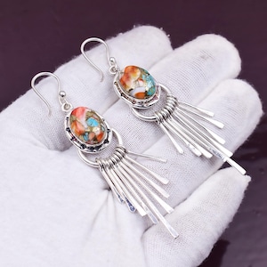 May include: A pair of silver earrings with oval-shaped, multicoloured stone settings. The stones feature orange, turquoise, and white hues. Below the stone, a series of silver fringe dangles. The earrings are displayed on a white surface.