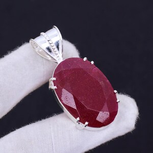 May include: A silver pendant featuring a large, oval-shaped ruby gemstone with a faceted cut. The pendant has a silver bail for attaching to a chain.