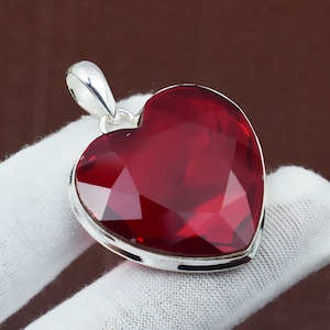 May include: A heart-shaped pendant with a faceted, deep red gemstone set in a silver-colored metal frame. The pendant has a loop at the top for a chain. The gemstone's cut creates many reflective surfaces.