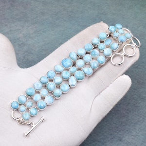 Könnte beinhalten: Ein silbernes Armband mit drei Reihen hellblauer, runder und ovaler Edelsteine. Das Armband hat einen Knebelverschluss. Die Edelsteine sind in einer Reihe angeordnet, wodurch ein geschichteter Effekt entsteht. Das Armband wird auf einer weißen Oberfläche präsentiert.