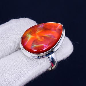 May include: A silver ring featuring a teardrop-shaped red opal gemstone. The opal displays a fiery iridescent sheen with flashes of green, yellow, and pink.