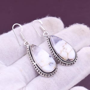 May include: A pair of silver-toned dangle earrings. Each earring features a teardrop-shaped white stone with gray and brown marbling, set in a detailed silver frame. The earrings have a hook closure.