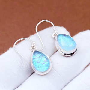 May include: Silver teardrop earrings featuring iridescent blue-green opal stones. The opals display a shimmering, multi-coloured effect, set in a polished silver bezel. The earrings are displayed on a white surface.
