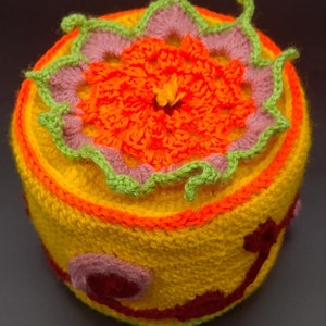 Puede incluir: Un gorro de crochet amarillo, naranja, rosa y verde con un diseño de flor en la parte superior.