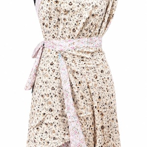 Könnte beinhalten: Ein schulterfreies Wickelkleid mit beiger Basis und dichtem braun-schwarzem Blumenmuster. Das Kleidungsstück verfügt über einen passenden Taillengürtel sowie einen kontrastierenden hellrosa und blauen Blumenbesatz an Ausschnitt, Taille und Saum.